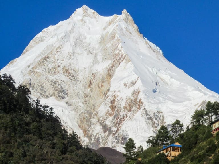 Trekking am Manaslu Circuit: tibetisch-nepalesischer Klosterkomplex an bewaldetem Hang mit pyramidalem Schneegipfel im Hintergrund.