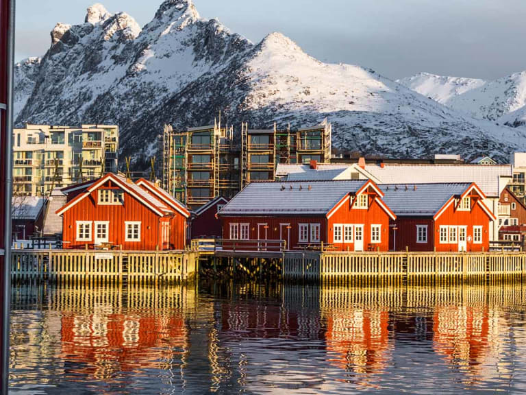 Rote Rorbu-Häuser in Svolvær spiegeln sich im Fjord, dahinter verschneite Berggipfel der Lofoten.