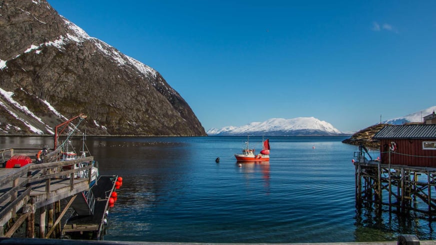 Bergwanderung in Nordnorwegen: Fischerhafen Koppangen mit rotem Bootshaus, Pier und schneebedeckten Bergen im Hintergrund.