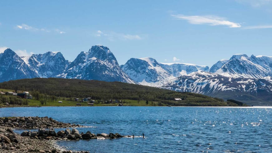 Bergwanderung in Norwegen: Blick über steiniges Fjordufer auf schneebedeckte Bergkette mit zentralem Gipfel im Frühsommer.