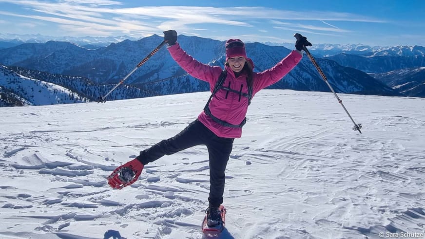 Schneeschuhwanderin in pinker Jacke posiert mit ausgebreiteten Armen auf schneebedecktem Gipfel am Spitzingsee.