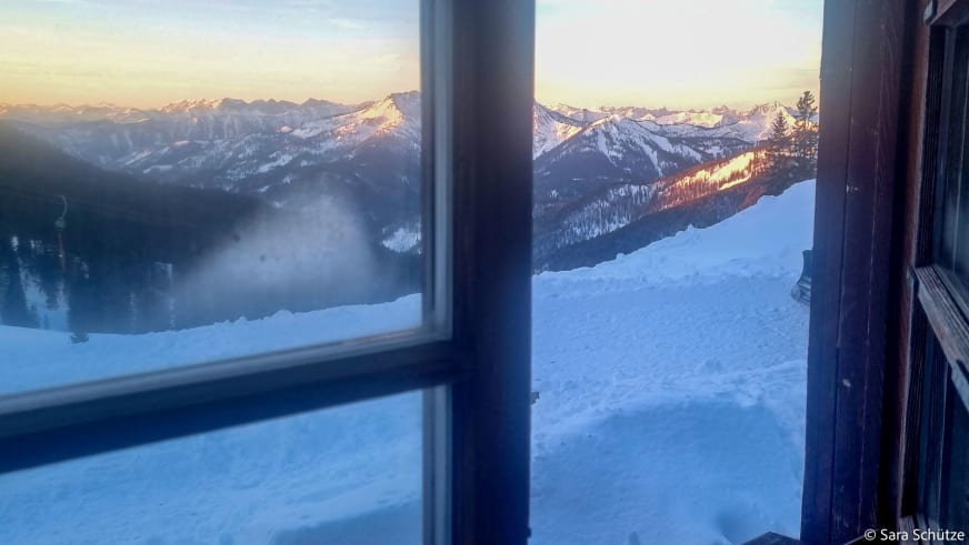 Schneeschuhtour am Spitzingsee: Blick durch ein Hüttenfenster auf verschneite Berghänge im Morgen- oder Abendlicht.