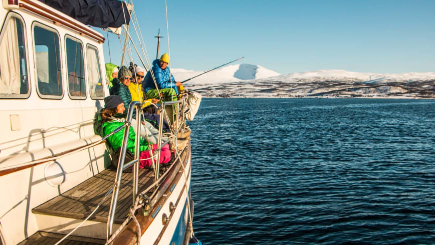 Skitour Norwegen: Fünf Skitourengeher angeln vom Achterdeck eines Boots auf dem Fjord vor schneebedeckten Bergen.