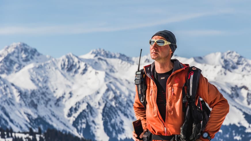 Skitour in Kirgistan: Bergführer in orangefarbener Jacke mit Rucksack vor schneebedeckten Gipfeln unter blauem Himmel.