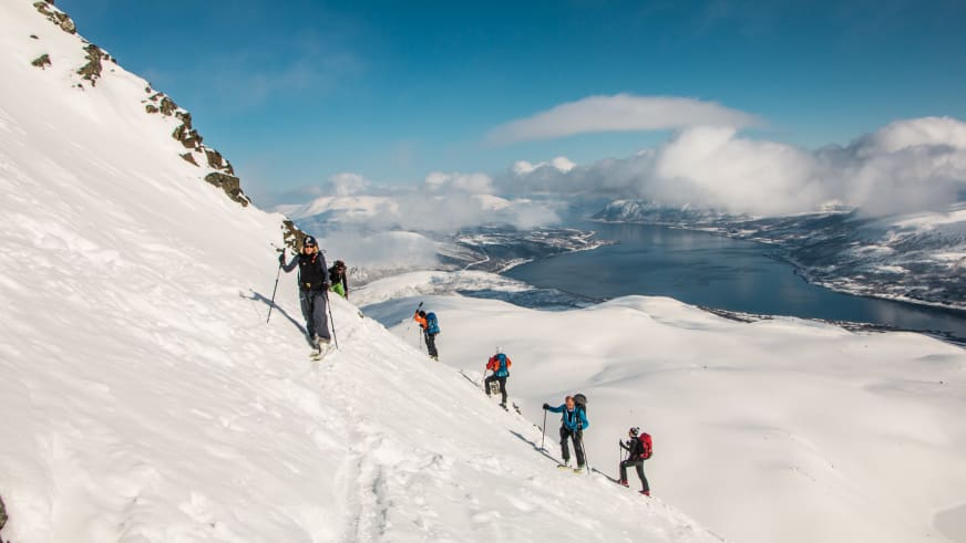 Fünf Skitourengeher steigen mit Fellen einen steilen verschneiten Hang über einem Fjord in Norwegen auf, Stützpunkt Schiff