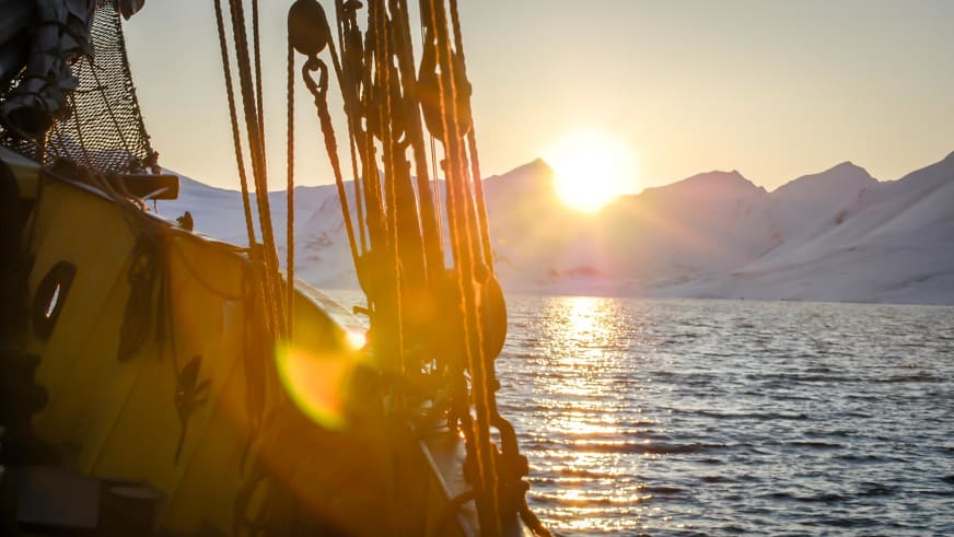 Skitour auf Spitzbergen: Blick vom Segelschiff auf verschneite Gipfel und tief stehende Sonne über dem Fjord.