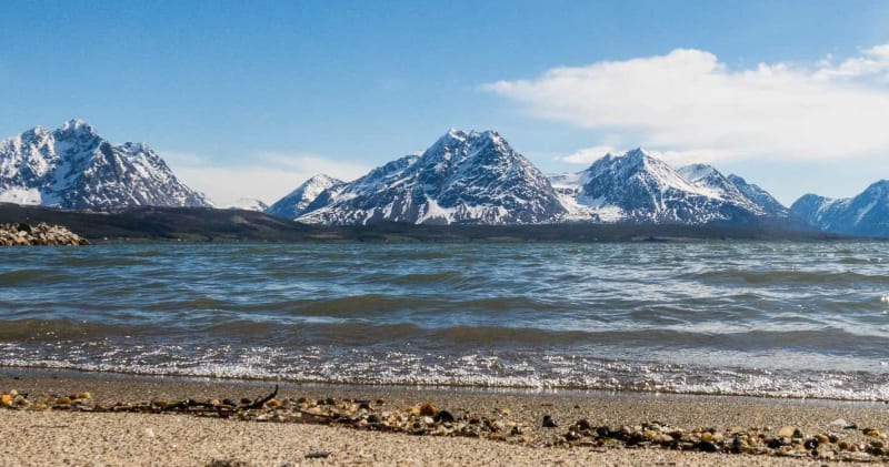 Bergwanderung in Lyngen: Kiesstrand am Fjord mit schneebedeckter Gebirgskette im Hintergrund.