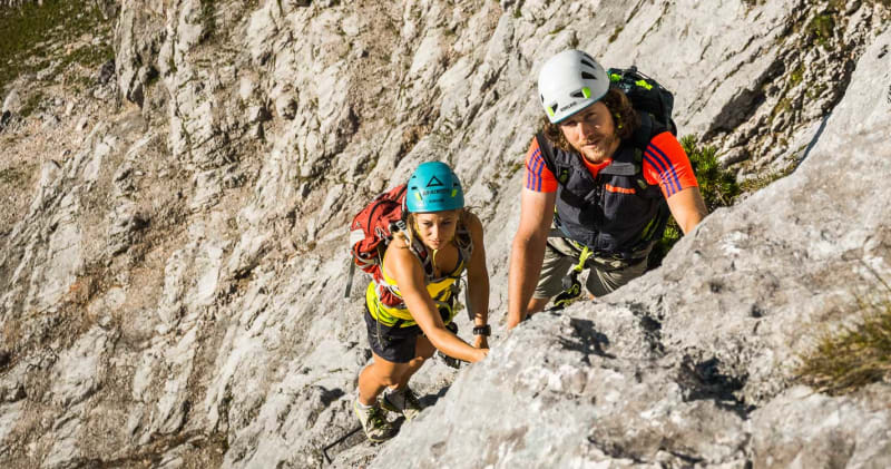 Klettersteig-Kurs: zwei Personen mit Helm klettern eine steile helle Kalkfelsplatte hinauf, gefuehrt von Alpine Welten.