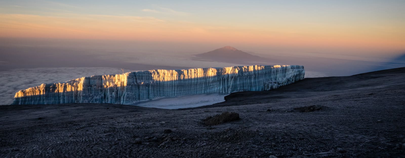 Expedition am Kilimanjaro: senkrechte Gletscherwand mit Eisschichtung über dunklem Vulkangestein bei Morgendämmerung.