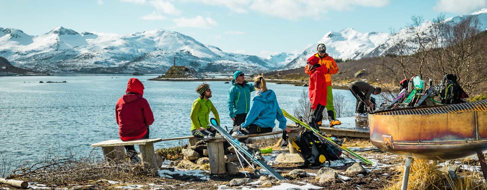 Gruppe von sieben Skitourengehern pausiert an einem Fjordufer auf den Lofoten mit schneebedeckten Bergen im Hintergrund.