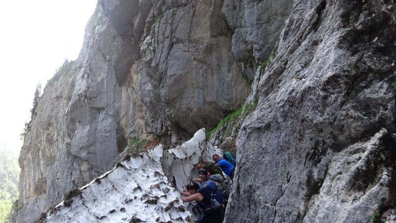 Drei Bergwanderer mit Rucksäcken traversieren einen Felsband-Weg über Schneefeld auf dem Weg zum Funtensee.