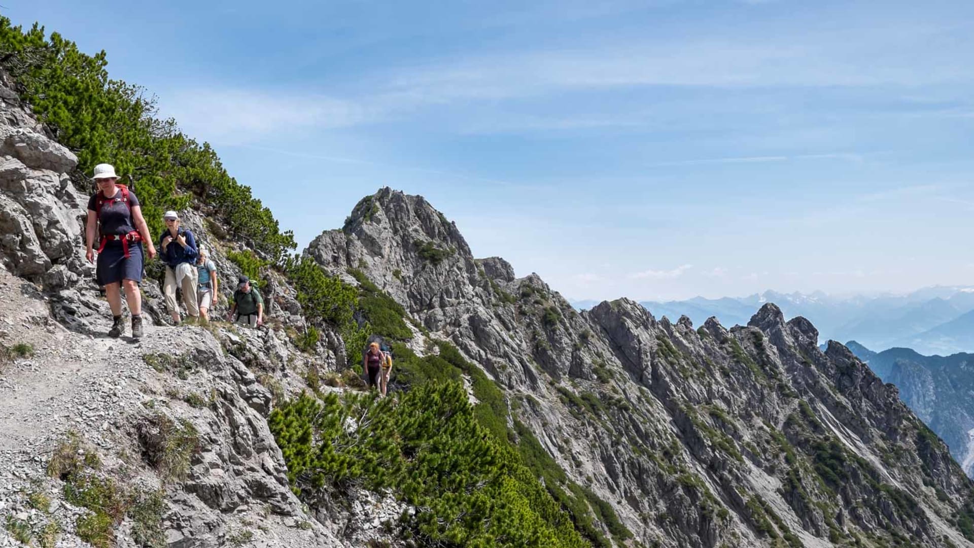 Fünf Bergwanderer traversieren einen Schotterweg entlang eines felsigen Grats mit Talblick in Liechtenstein.