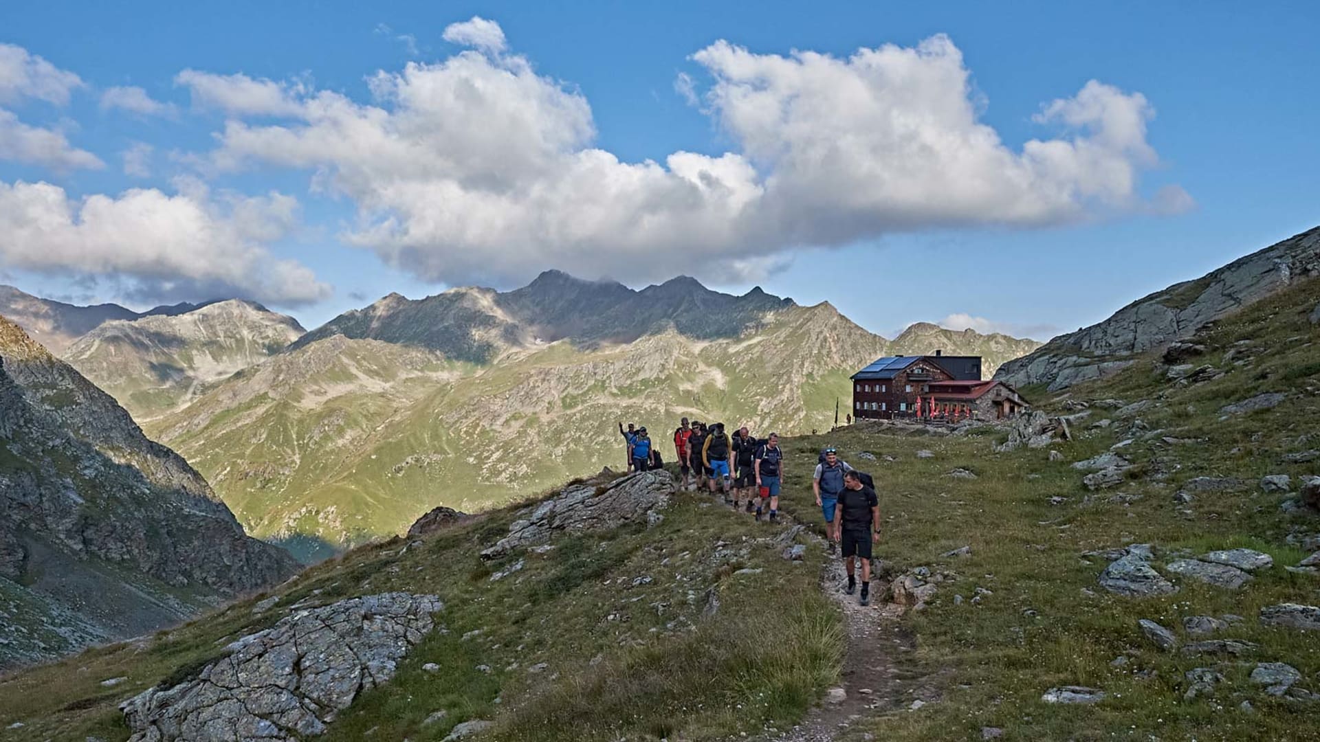 Bergwanderung im Verwall: Gruppe von Bergwanderern auf schmalem Pfad über Bergrücken mit Berghütte im Mittelgrund.
