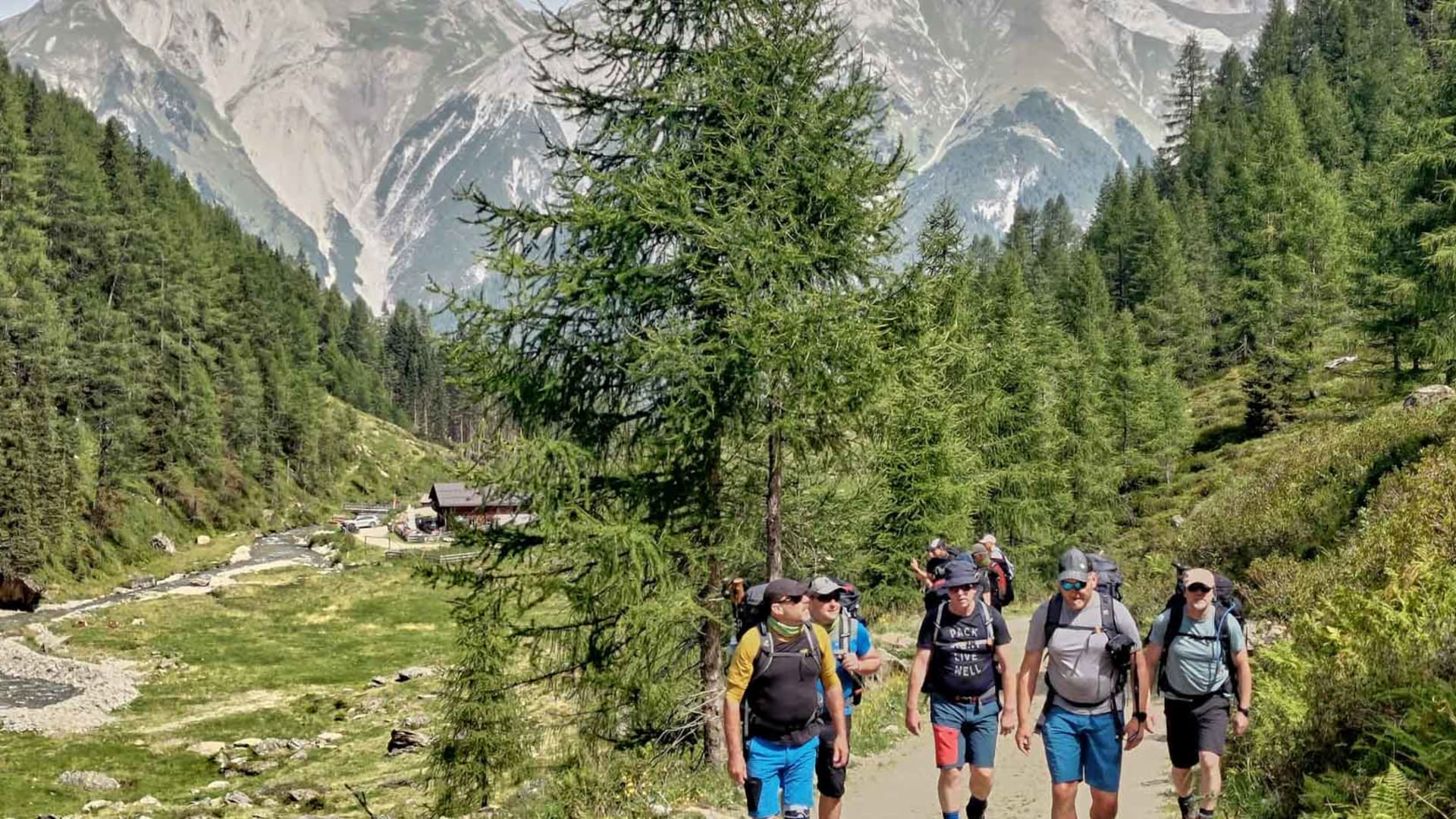Bergwanderung im Verwall: Gruppe von Bergwanderern mit Rucksäcken auf breitem Schotterweg entlang eines Bachbetts.
