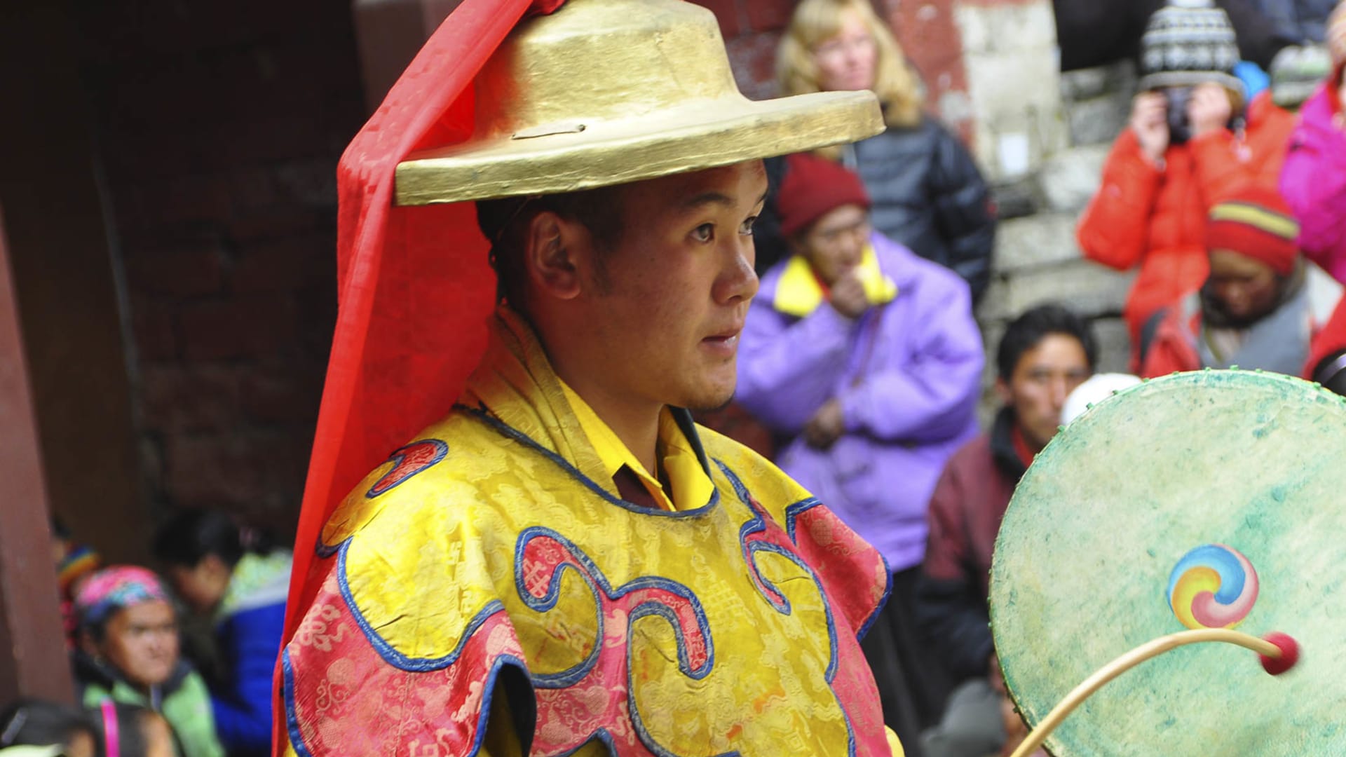 Klosterfestival im Kloster Tengboche: Zeremonienträger in gelbem Brokatgewand mit Trommel vor bunter Zuschauermenge in Nepal.