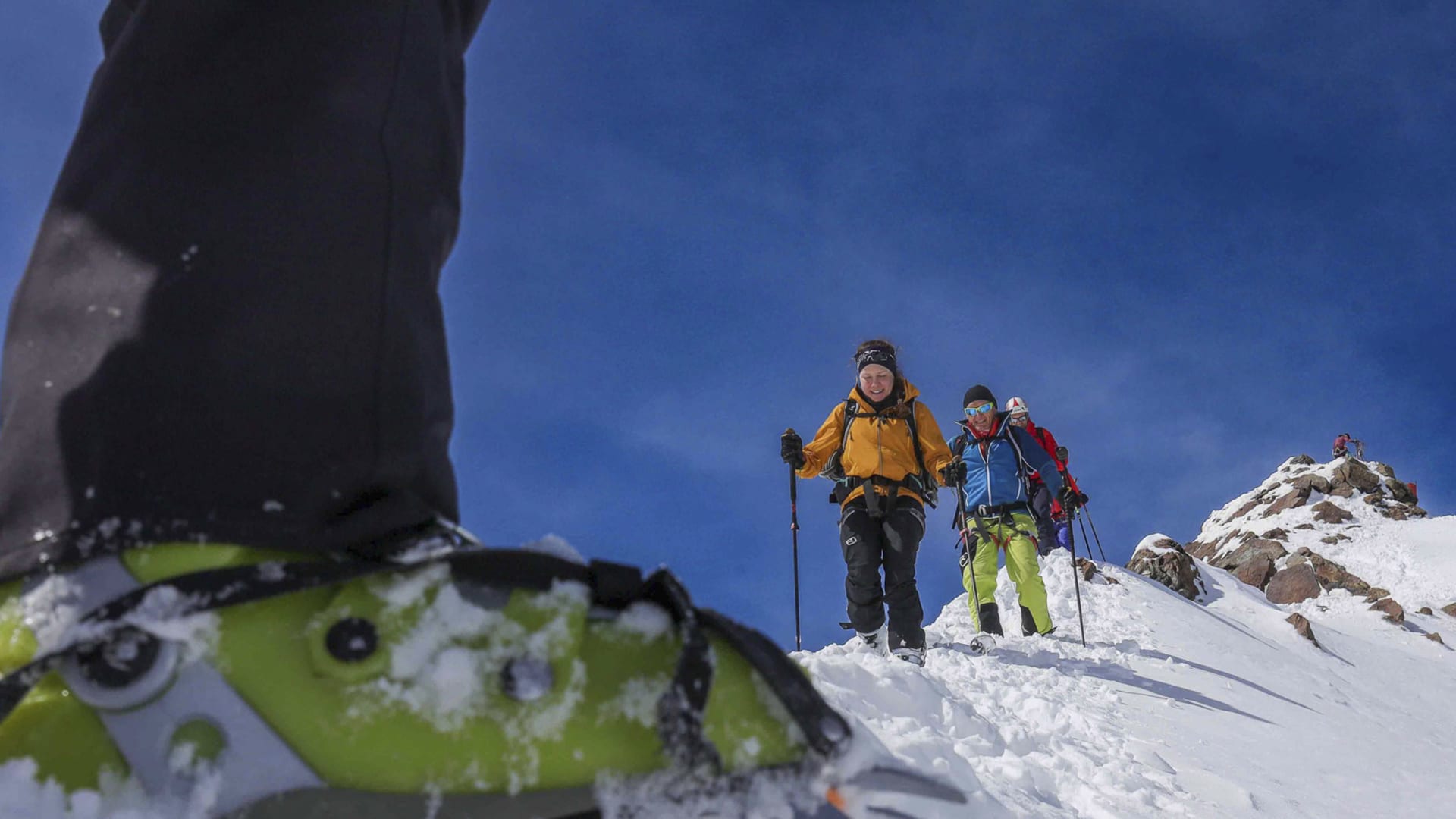 Zwei Skitourengeher steigen mit Stöcken die Zufallspitze hinauf, im Vordergrund ein Steigeisen im Schnee, blauer Himmel.