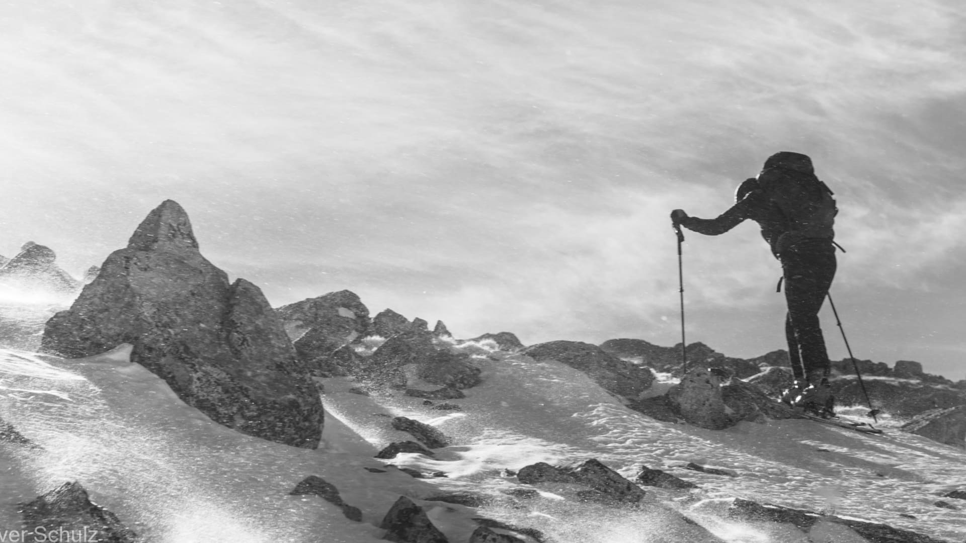 Skitouren-Geher mit Rucksack auf felsigem, schneebedecktem Grat im Engadin, aufgewirbelter Schnee und dichte Wolken