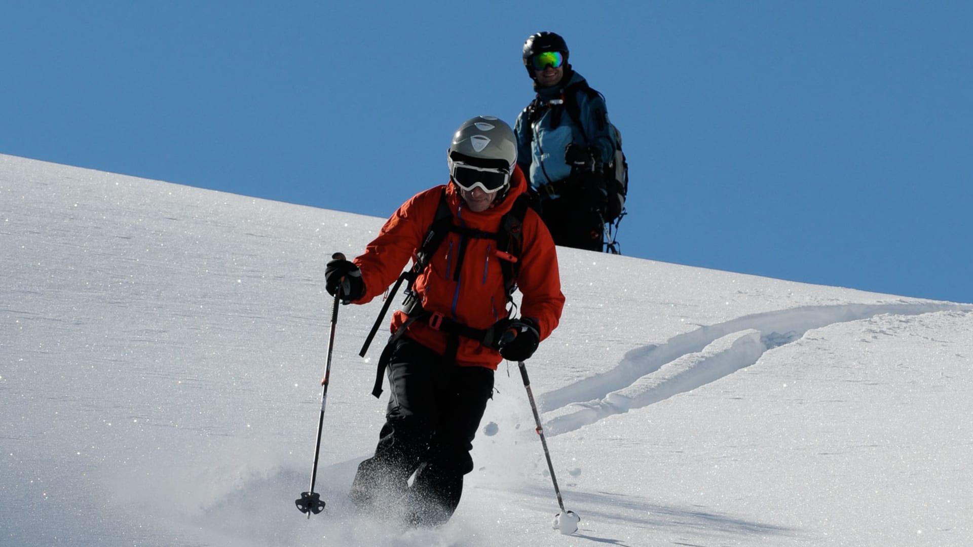 Zwei Personen üben Tiefschneetechnik auf einer Skitour an einem unverspurten Steilhang mit Pulverschnee unter blauem Himmel.