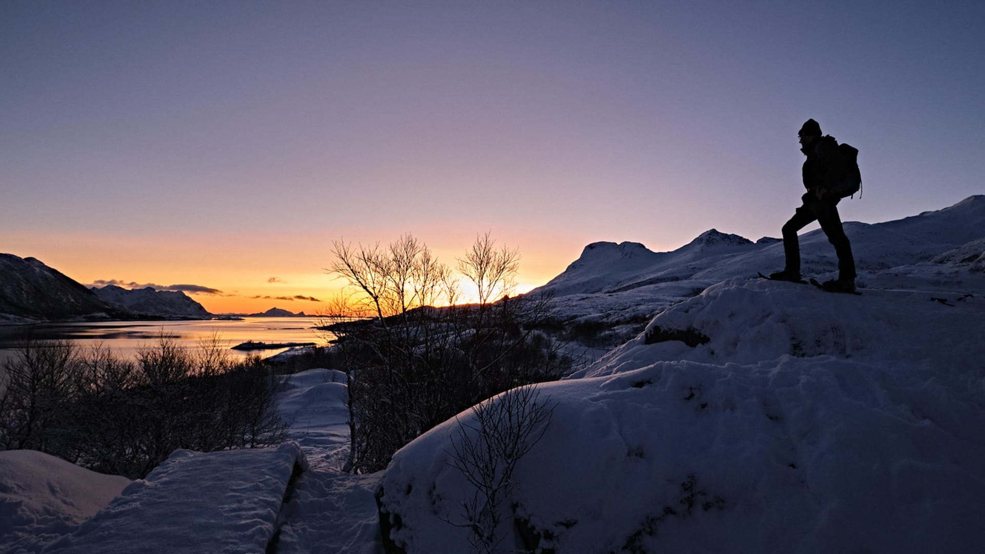 Winterwanderer als Silhouette auf einem Felsvorsprung blickt bei Sonnenuntergang über einen Fjord auf den Lofoten.