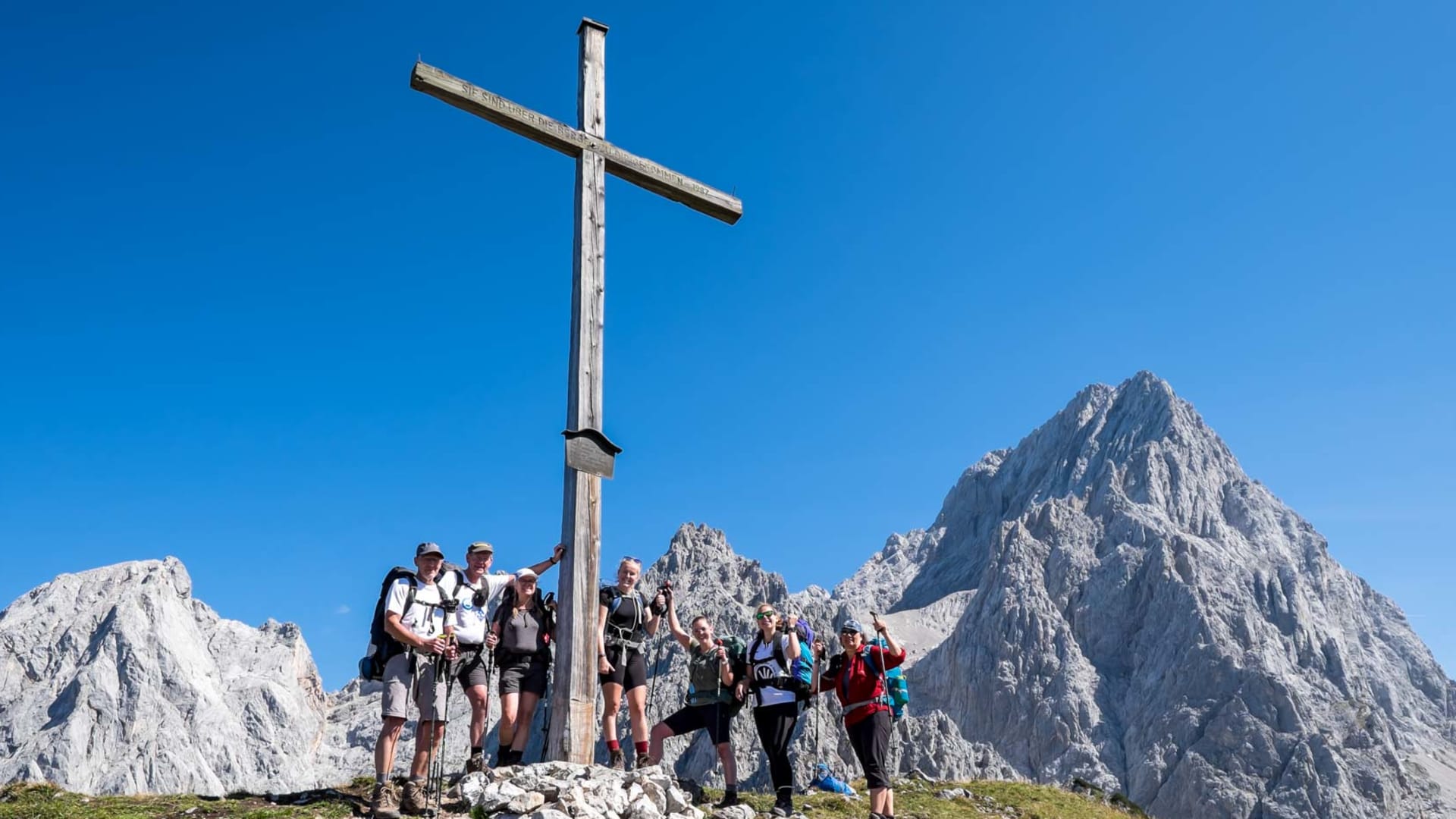 Sechs Bergwanderer posieren am Holzgipfelkreuz des Hohen Dachsteins mit Kalkfelsgipfeln im Hintergrund.