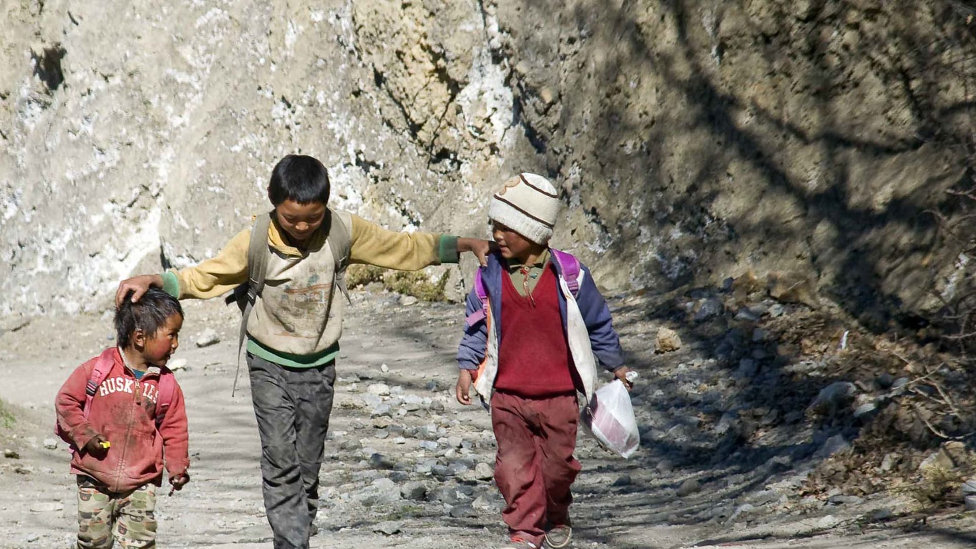 Trekking in Nepal: drei Kinder laufen auf einem steinigen Pfad entlang einer Felswand in einer engen Schlucht.