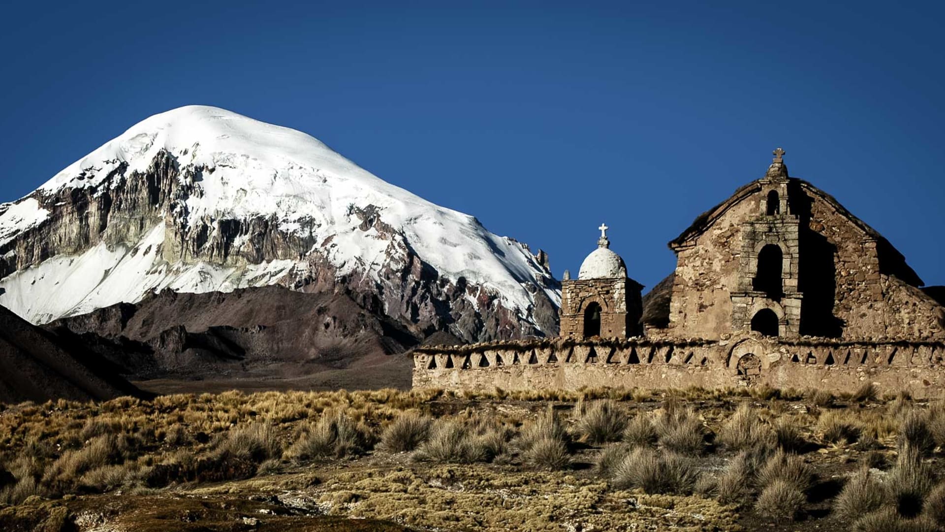 Expedition auf den Sajama: schneebedeckter Vulkankegel mit kolonialer Steinkirche im Vordergrund auf dem bolivianischen Hochplateau.