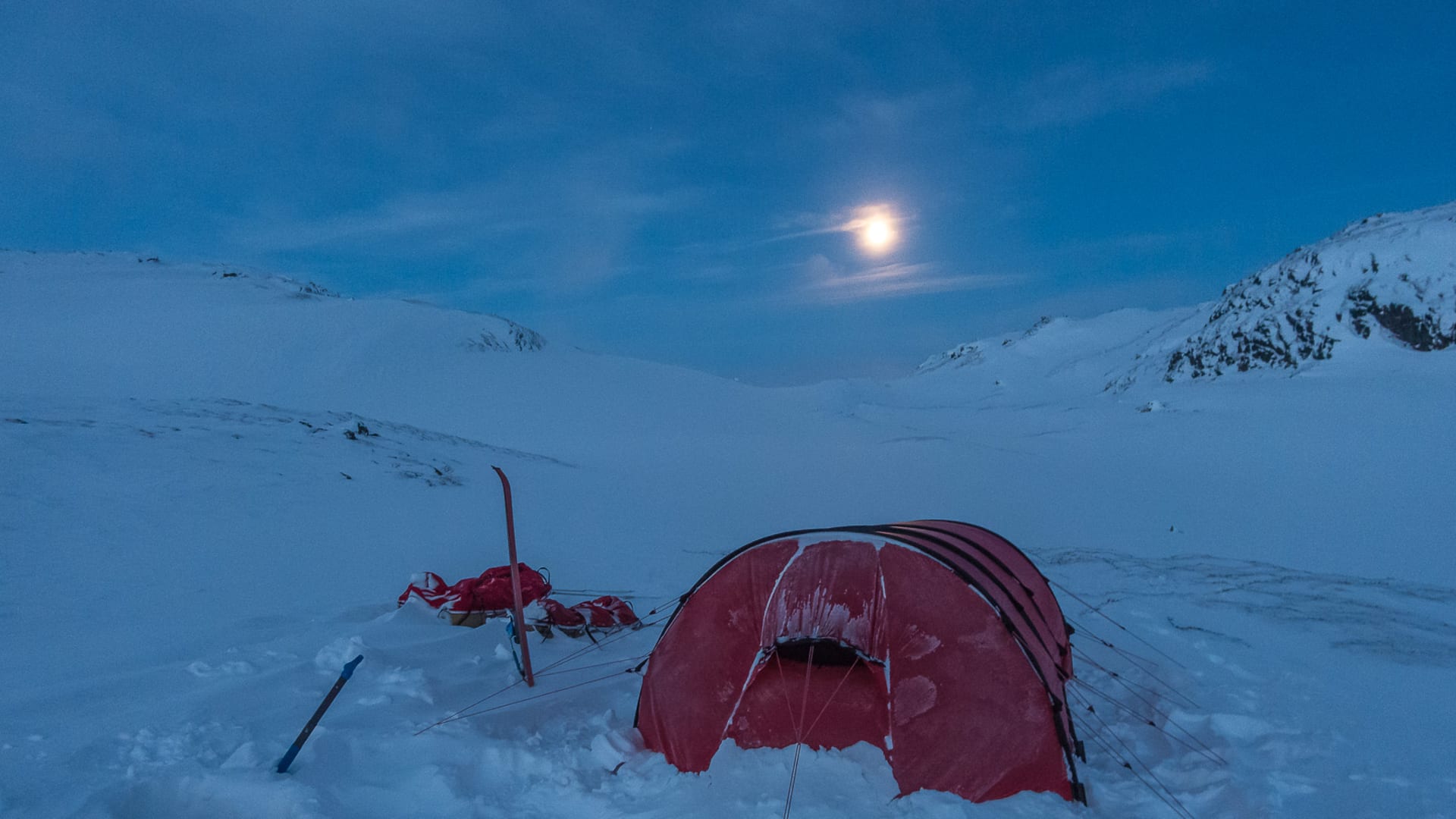 Expedition im Sarek: rotes Zelt auf Schneeplateau mit Tourenskiern bei blauer Stunde und Vollmond über Bergkamm.