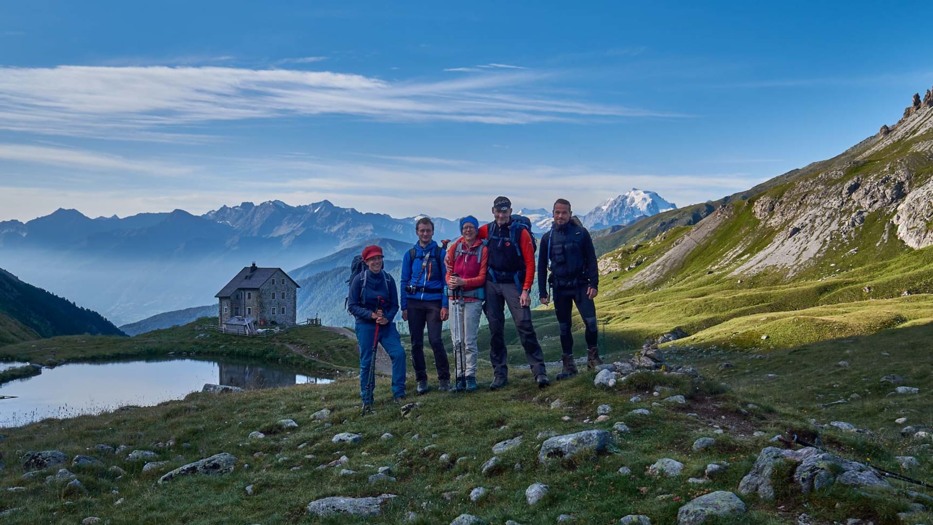 Fünf Bergwanderer posieren auf einer Hochalm-Kuppe im Engadin mit Alpensee und Berghütte im Hintergrund.