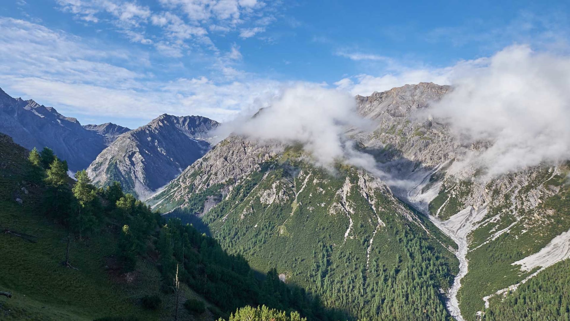 Panoramablick ins bewaldete Alpental im Engadin mit felsigen Gipfeln und Wolken aus erhöhter Perspektive.