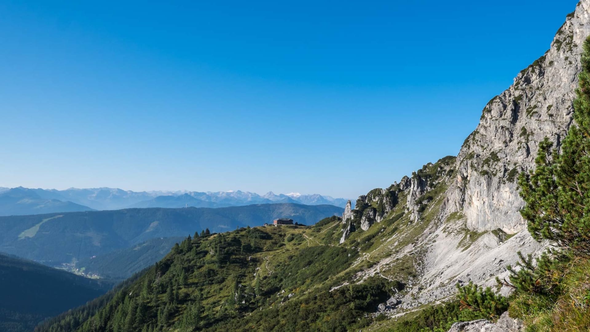 Panoramablick auf eine Berghütte am bewaldeten Rücken des Hohen Dachstein mit Talblick und tiefblauem Himmel.