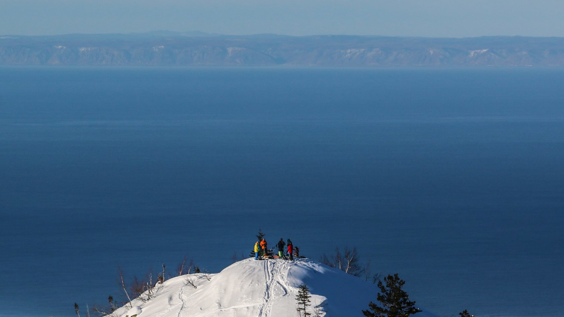Gruppe von Skitourengehern pausiert auf einem schneebedeckten Gipfel mit Blick auf den tiefblauen Baikalsee in Sibirien.
