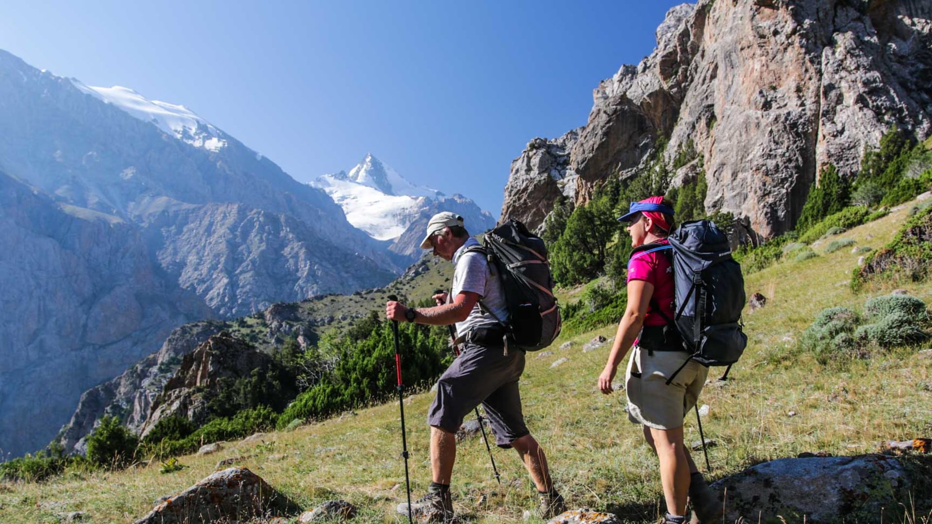 Zwei Trekker mit Rucksäcken und Teleskopstöcken steigen einen grasig-felsigen Hang in Kirgistan auf.