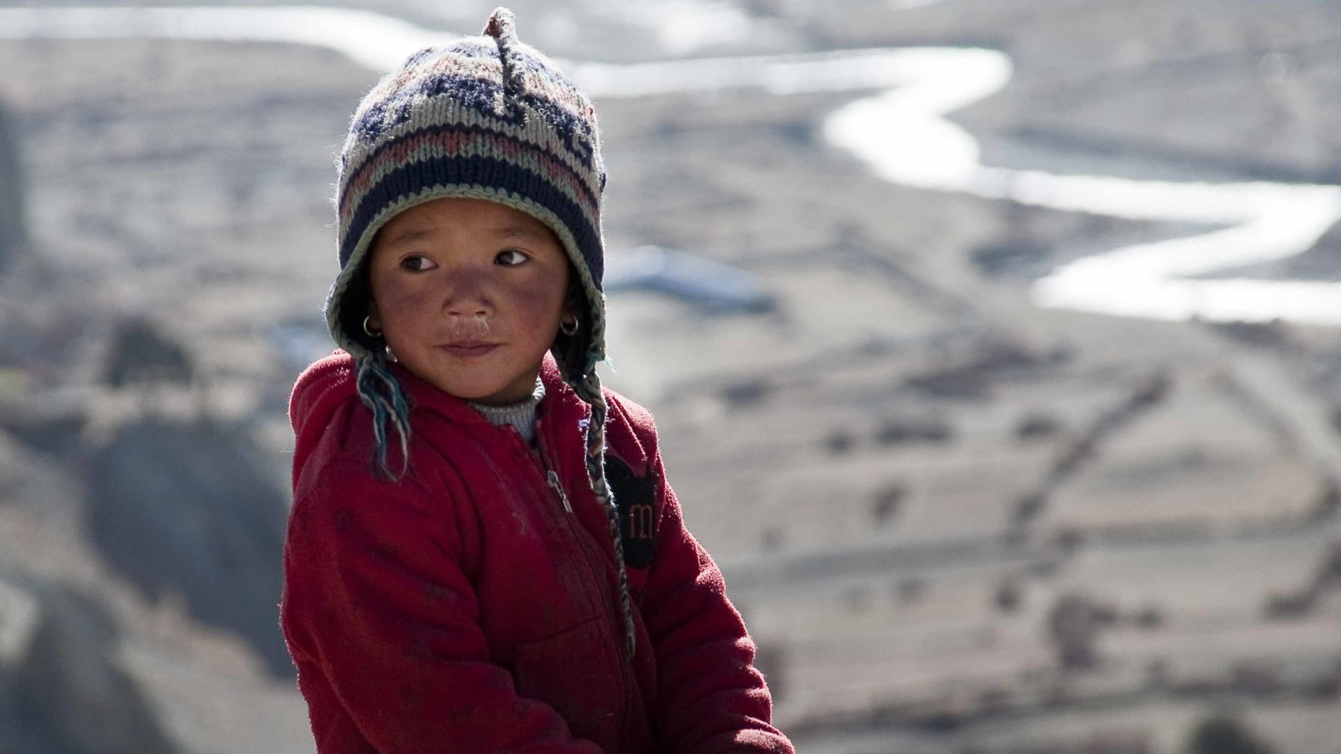 Trekking Nepal: Nahporträt eines Kindes mit Wollmütze und roter Jacke vor trockener Hochgebirgslandschaft im Himalaya.