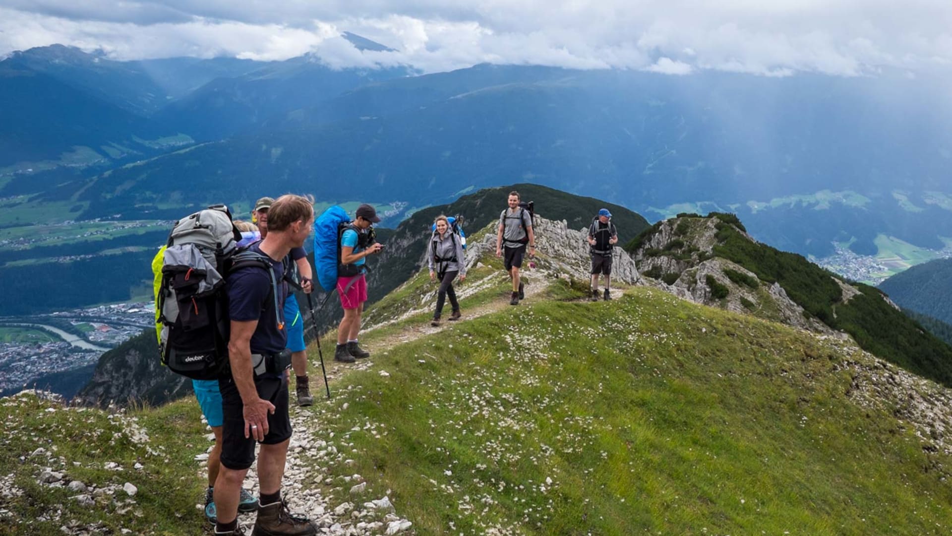 Sechs Bergwanderer mit Rucksäcken gehen auf schmalem Grasgrat am Karwendel Höhenweg mit Talblick.