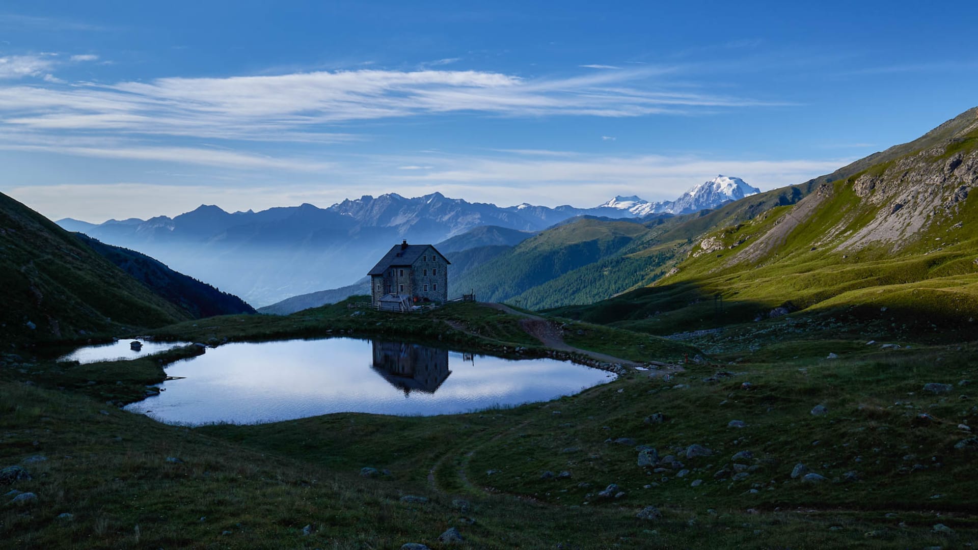 Berghütte im Engadin mit herzförmigem Bergsee im Vordergrund zur Blauen Stunde, schneebedeckte Gipfel im Hintergrund.