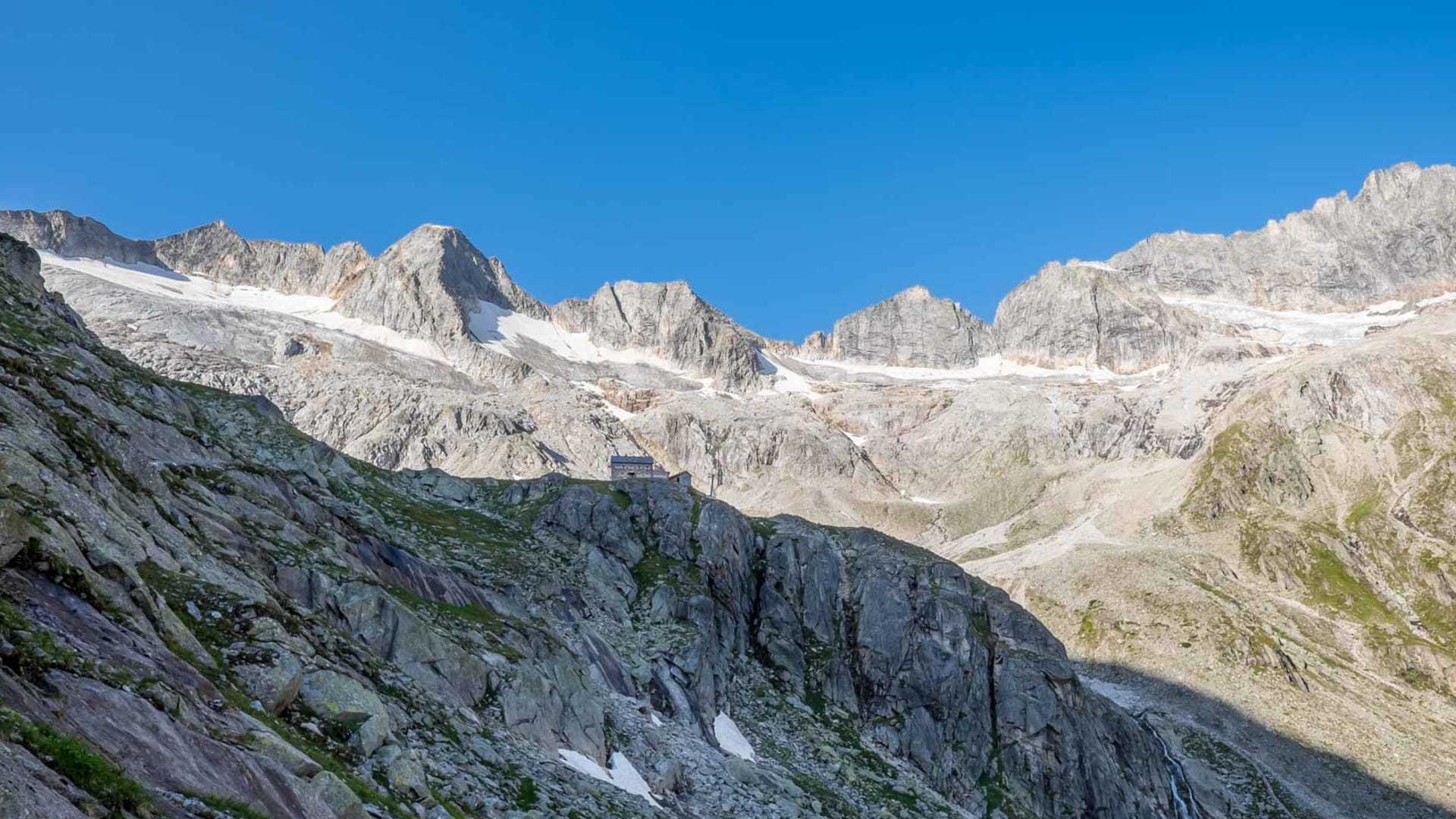 Bergwanderung im Zillertal: Richterhütte auf Felsvorsprung vor breitem Bergmassiv mit Gletscherfeldern und blauem Himmel.
