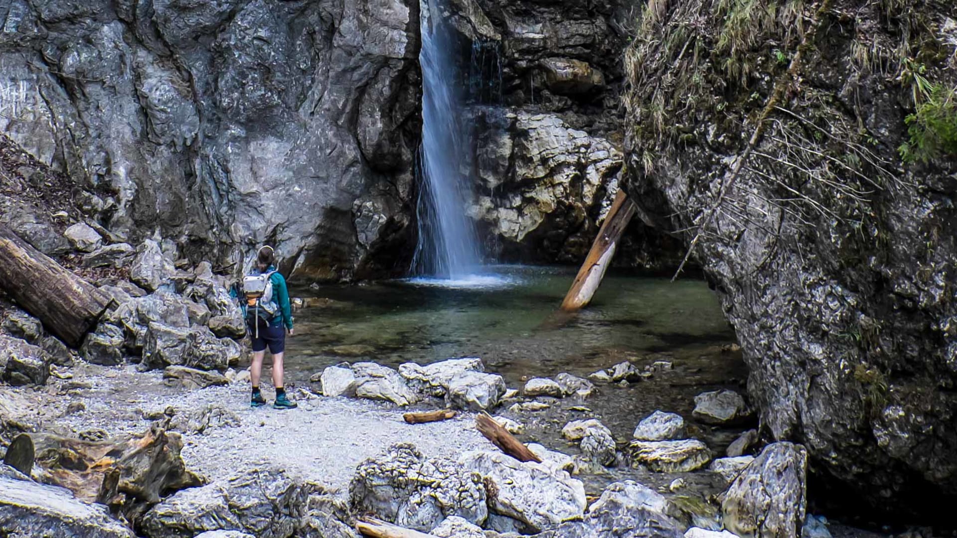 Bergwanderer steht in einer Kalksteinschlucht der Kocheler Berge vor einem Wasserfall mit türkisgrünem Becken.