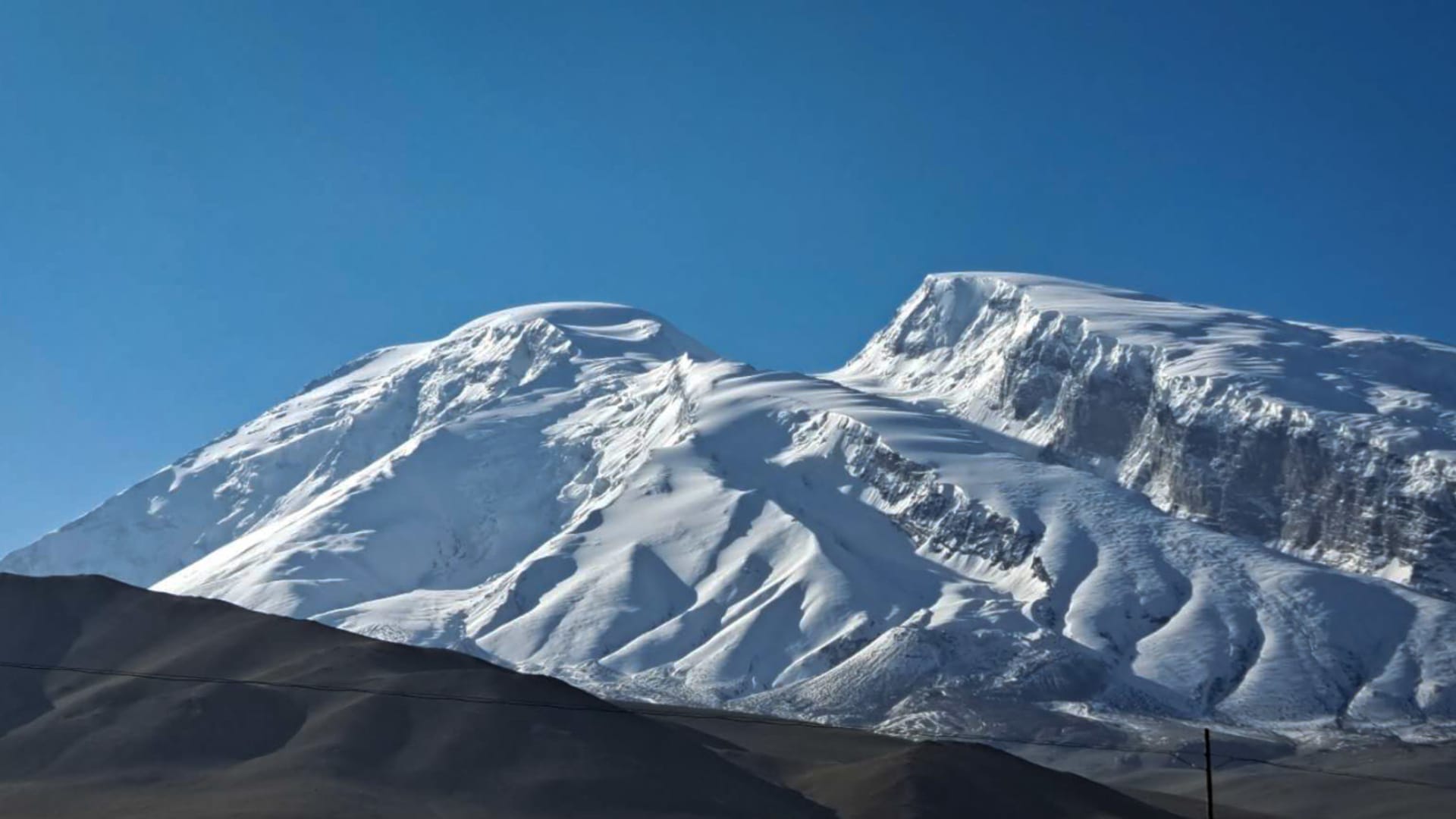 Expedition zum Muztagh Ata: schneebedecktes Doppelgipfelmassiv mit Gletscherhängen und dunklem Vordergrundgelände