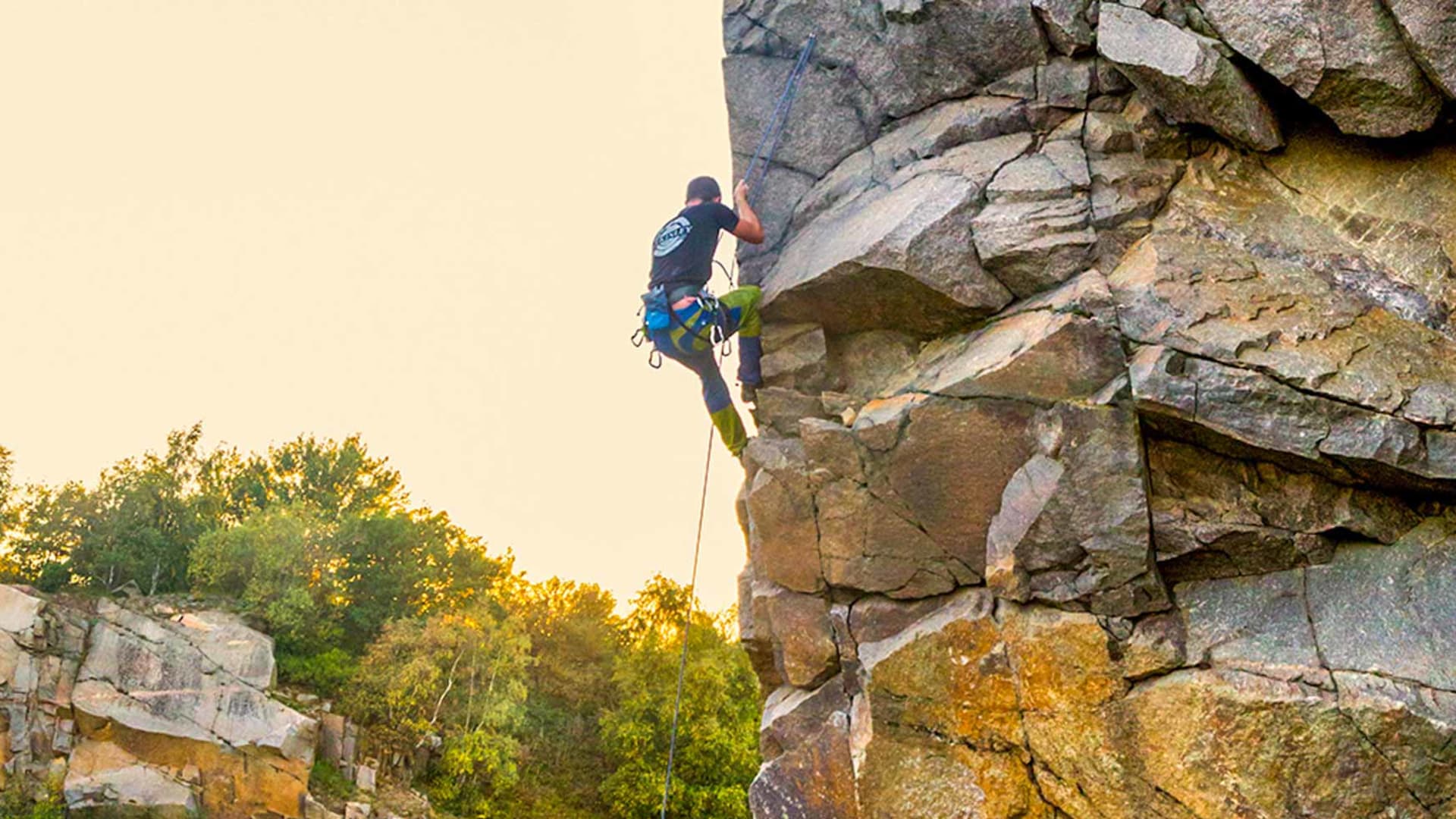 Kletterer mit Gurt und blauem Seil steigt eine senkrechte Granitwand auf Bornholm im Abendlicht auf.
