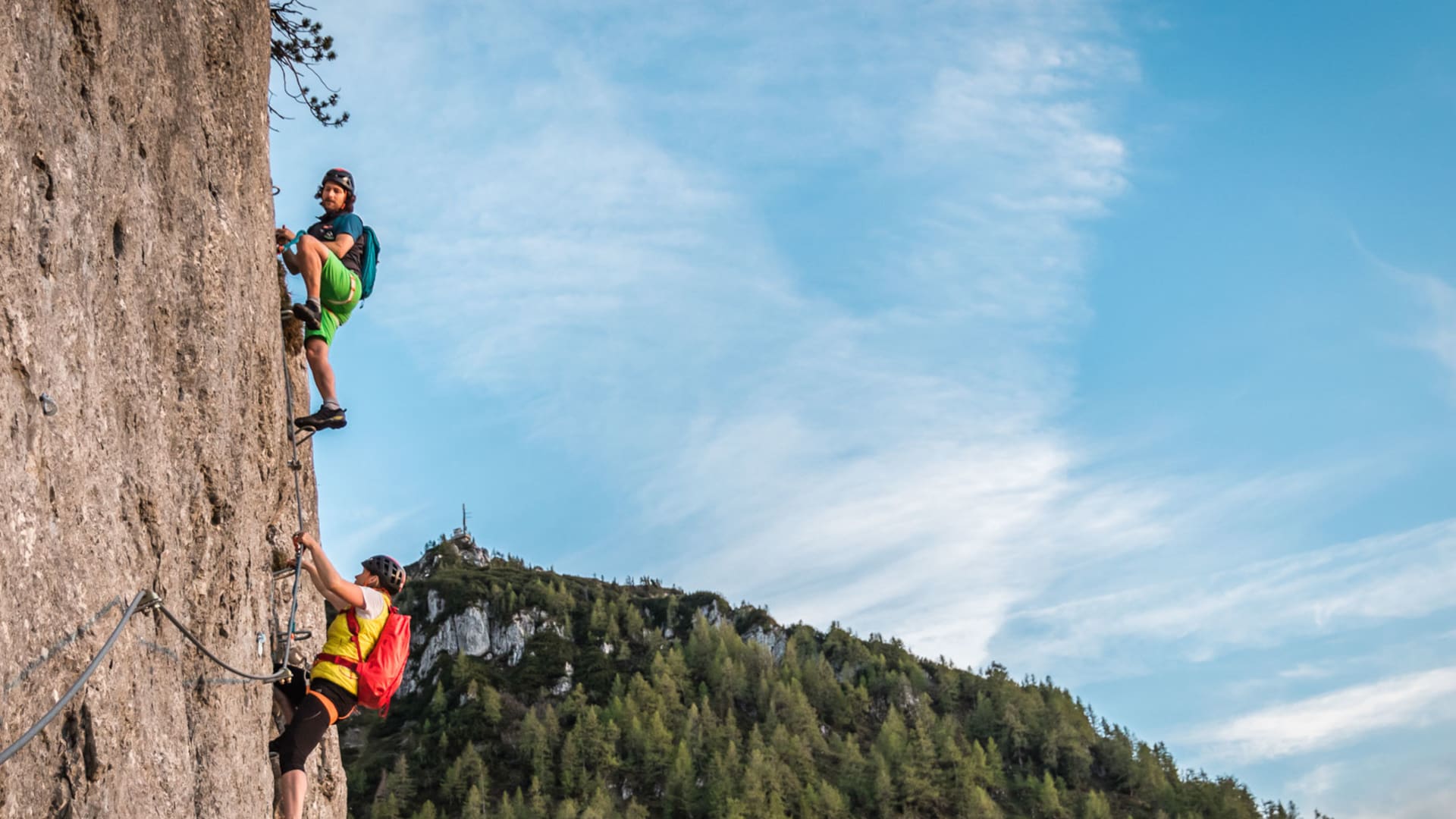 Zwei Klettersteiggeher klettern am Jenner eine senkrechte Kalksteinwand auf, im Hintergrund Nadelwald und Bergkuppe.
