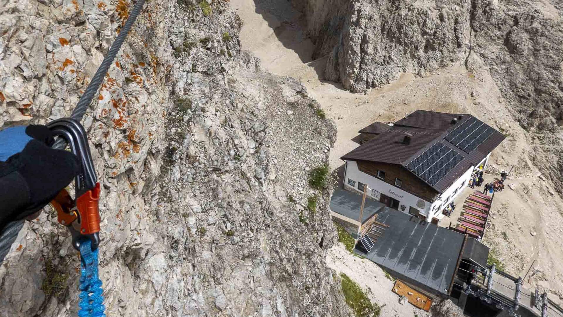 Blick vom Klettersteig am Langkofel senkrecht nach unten auf eine Berghütte mit Terrasse im Sella-Gebiet der Dolomiten.