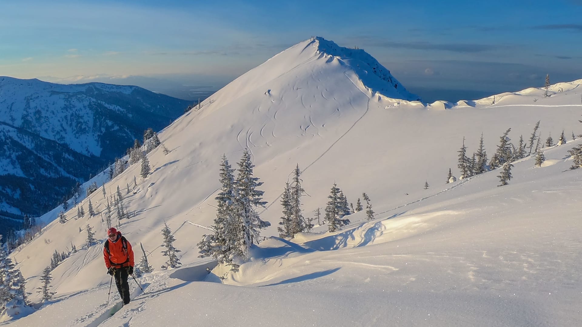 Skitourengeher mit roter Jacke und Rucksack steigt am Baikalsee einen verschneiten Hang zum Gipfel auf.