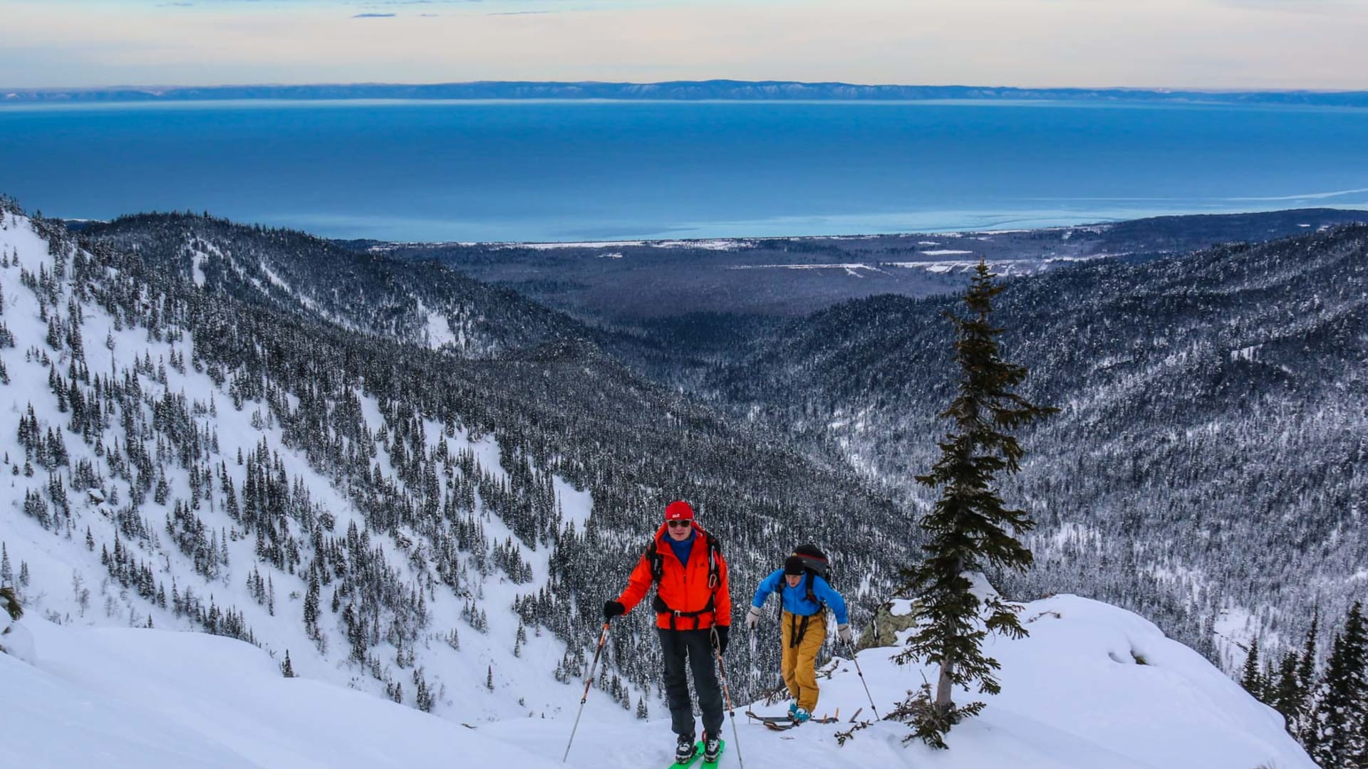 Zwei Skitourengeher steigen mit Tourenski und Fellen einen verschneiten Hang auf, im Hintergrund der Baikalsee in Sibirien.
