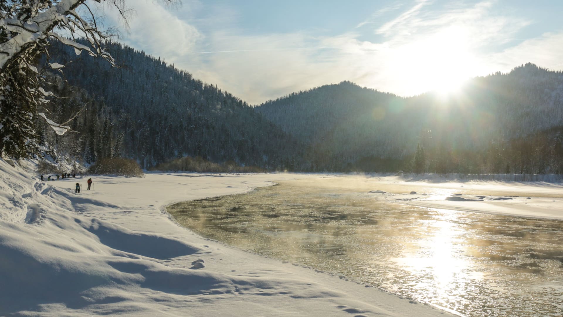 Skitourengeher am teilweise zugefrorenen Fluss in Sibirien mit schneebedeckten Nadelbäumen im Gegenlicht der Wintersonne.