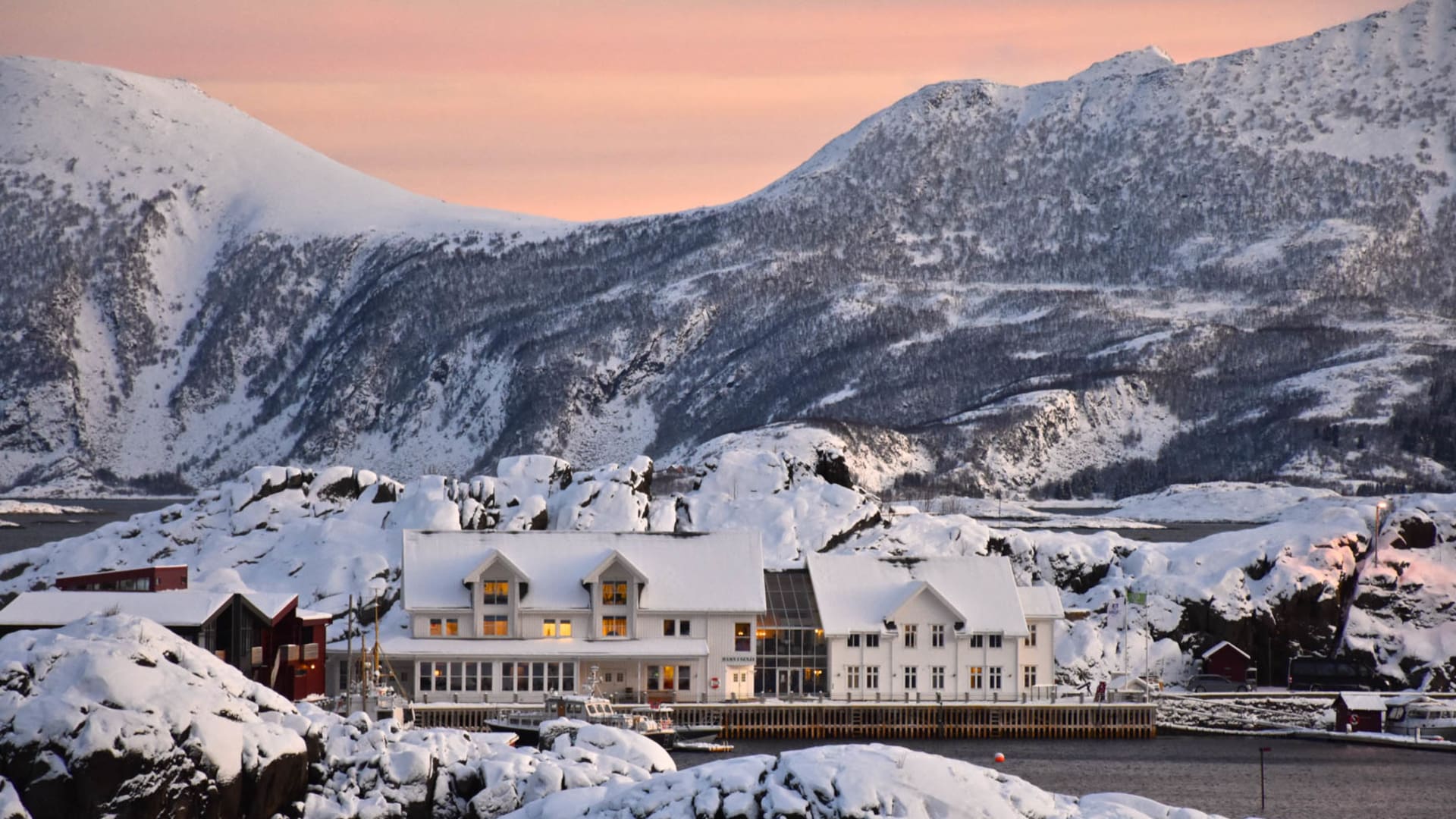 Skitour Senja Norwegen: Gebäudekomplex Hamn i Senja am verschneiten Ufer mit steilen schneebedeckten Bergen im Hintergrundund