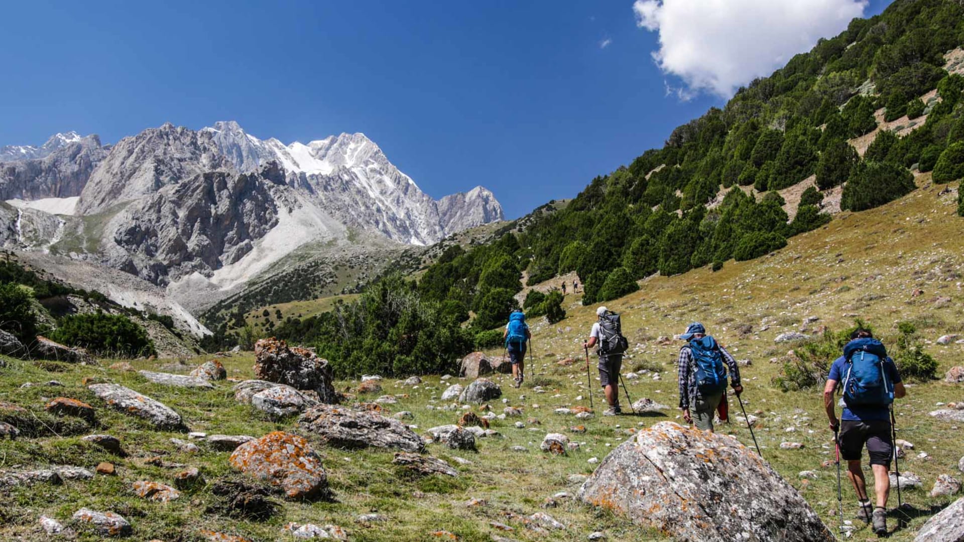 Vier Trekker mit großen Rucksäcken und Wanderstöcken steigen einen felsigen Hang im Pamir in Kirgistan auf.