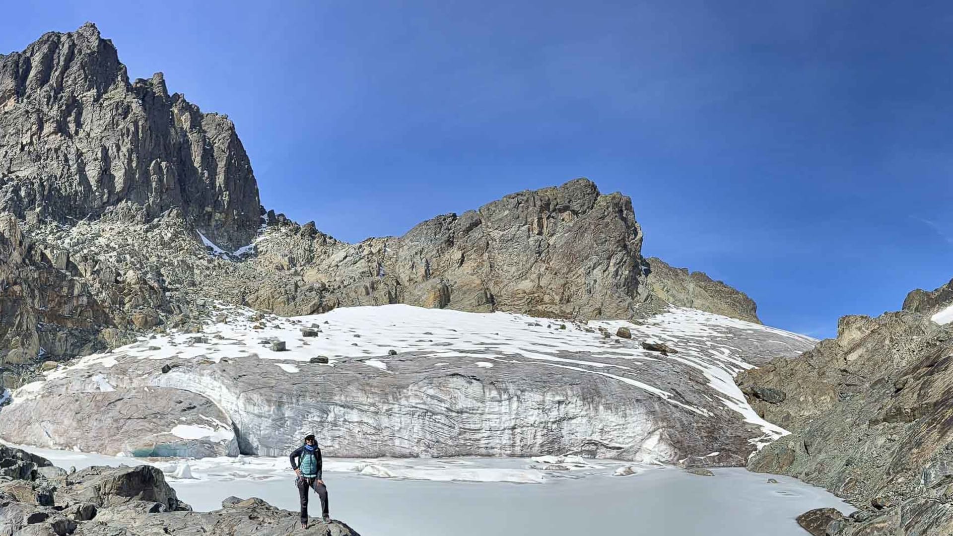 Trekker steht auf einem Felsblock an einem Gletschersee mit Eisfeld und Felsformationen im Ruwenzori Gebirge Uganda.