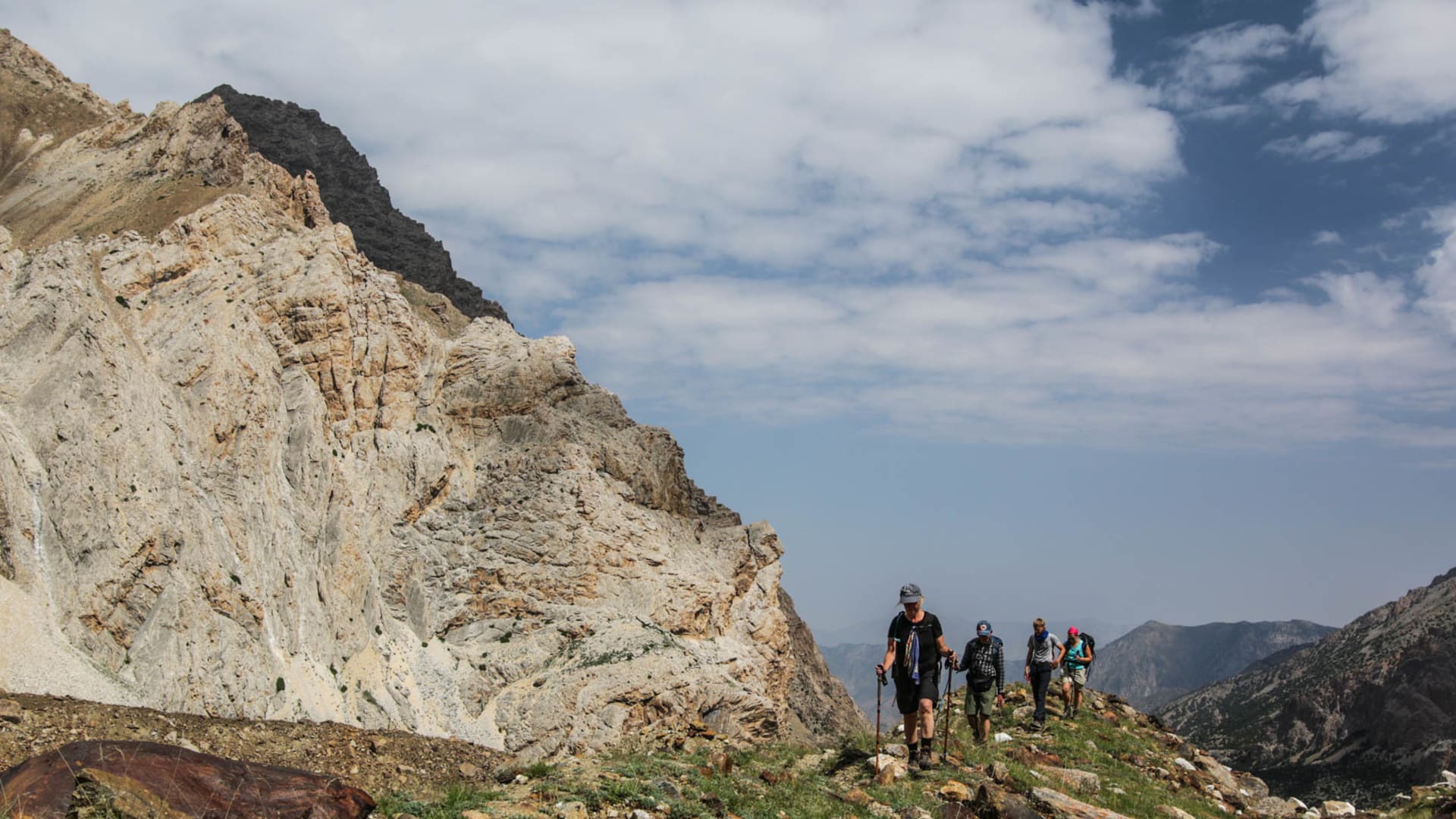 Vier Trekker mit Rucksäcken und Wanderstöcken auf einem grasigen Hang neben einer hellen Kalksteinwand in Kirgistan.