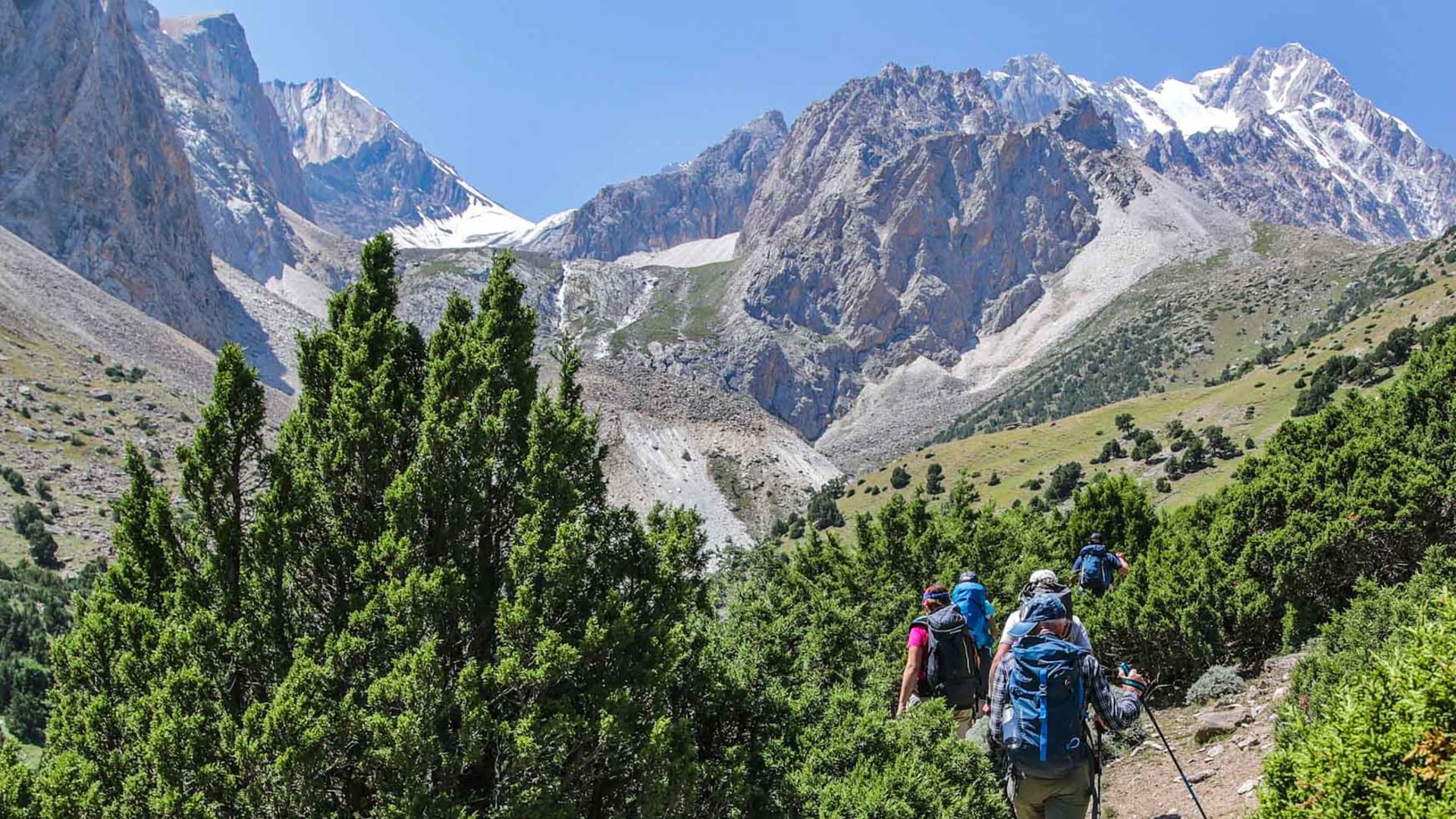 Trekking-Gruppe mit großen Rucksäcken und Wanderstöcken steigt auf schmalem Erdpfad durch ein felsiges Hochgebirgstal in Kirgistan