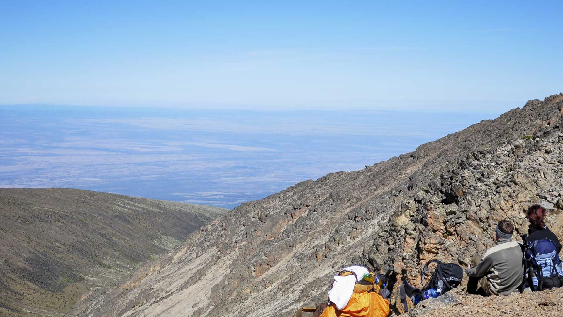 Zwei Trekker pausieren sitzend am felsigen Hang des Mount Kenia mit Blick auf die weite Tiefebene Kenias.